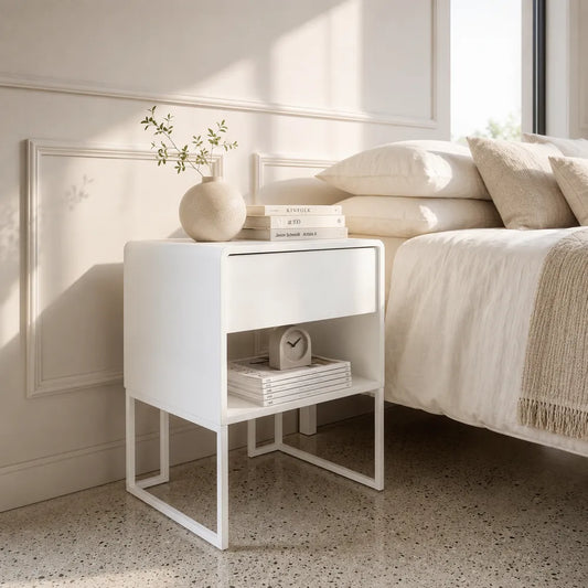 A modern white bedside table with a single drawer and open lower shelf, styled next to a bed in a bright, neutral bedroom.