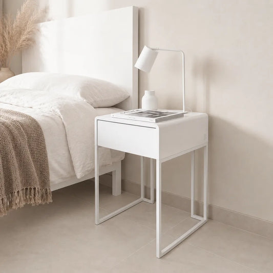 A modern white nightstand with a single drawer and open metal legs, styled next to a bed in a minimalist bedroom setting.