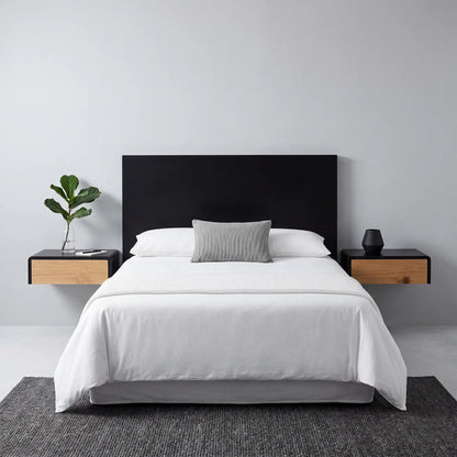A modern bed setup with a black headboard, white bedding, a gray accent pillow, and two floating wooden bedside tables against a light gray wall.