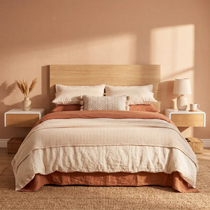 A neatly made bed in a warm, minimalist bedroom. The bedding set features a textured cream quilt, rust-orange sheets and duvet, and a patterned beige accent pillow.
