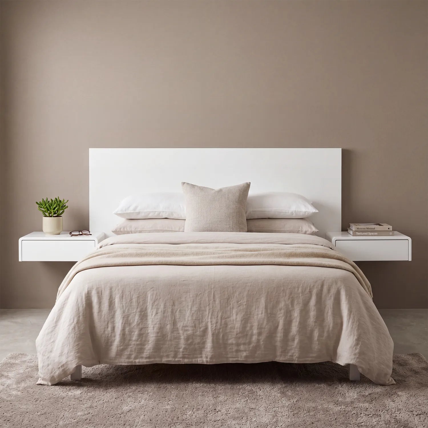 A minimalist bed setup in a neutral bedroom, featuring a white rectangular headboard, light beige linen bedding, matching pillows, and two floating white side tables with a small potted plant and books against a taupe wall.