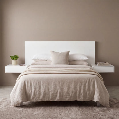 A minimalist bed setup in a neutral bedroom, featuring a white rectangular headboard, light beige linen bedding, matching pillows, and two floating white side tables with a small potted plant and books against a taupe wall.