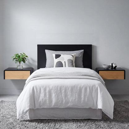 A neatly made bed with white bedding and a gray patterned accent pillow, centered against a light gray wall. The bed has a black upholstered headboard and is flanked by two floating bedside tables, one with a green plant and the other with small decor items.
