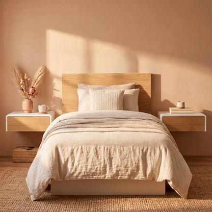 A neatly made bed with cream bedding and textured pillows, set against a warm beige wall with floating white and wood nightstands on each side, styled with a vase of dried grasses and a small cup.