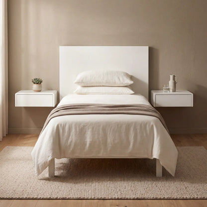 A minimalist single bed with a white headboard and light beige bedding, styled in a neutral bedroom setting with two floating white side tables, a textured rug, and a warm beige wall.