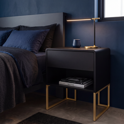 A modern black nightstand with gold metal legs and an open shelf, styled next to a bed in a dark blue bedroom. A slim gold table lamp sits on top of the nightstand, which also holds a small blue ceramic vase and books.