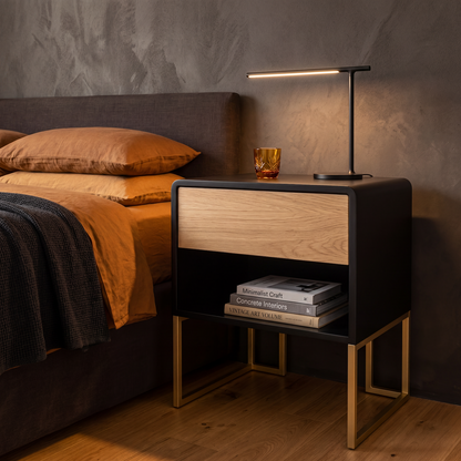 A modern black nightstand with a light oak wood drawer front and gold metal legs, styled next to a bed in a bedroom setting with a black table lamp, books, and an amber glass on top.