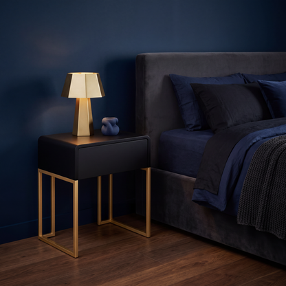 A modern bedside table with a black top and gold metal legs, styled next to a bed with dark blue bedding in a dark blue room. A lit geometric table lamp and a small blue vase sit on the table.