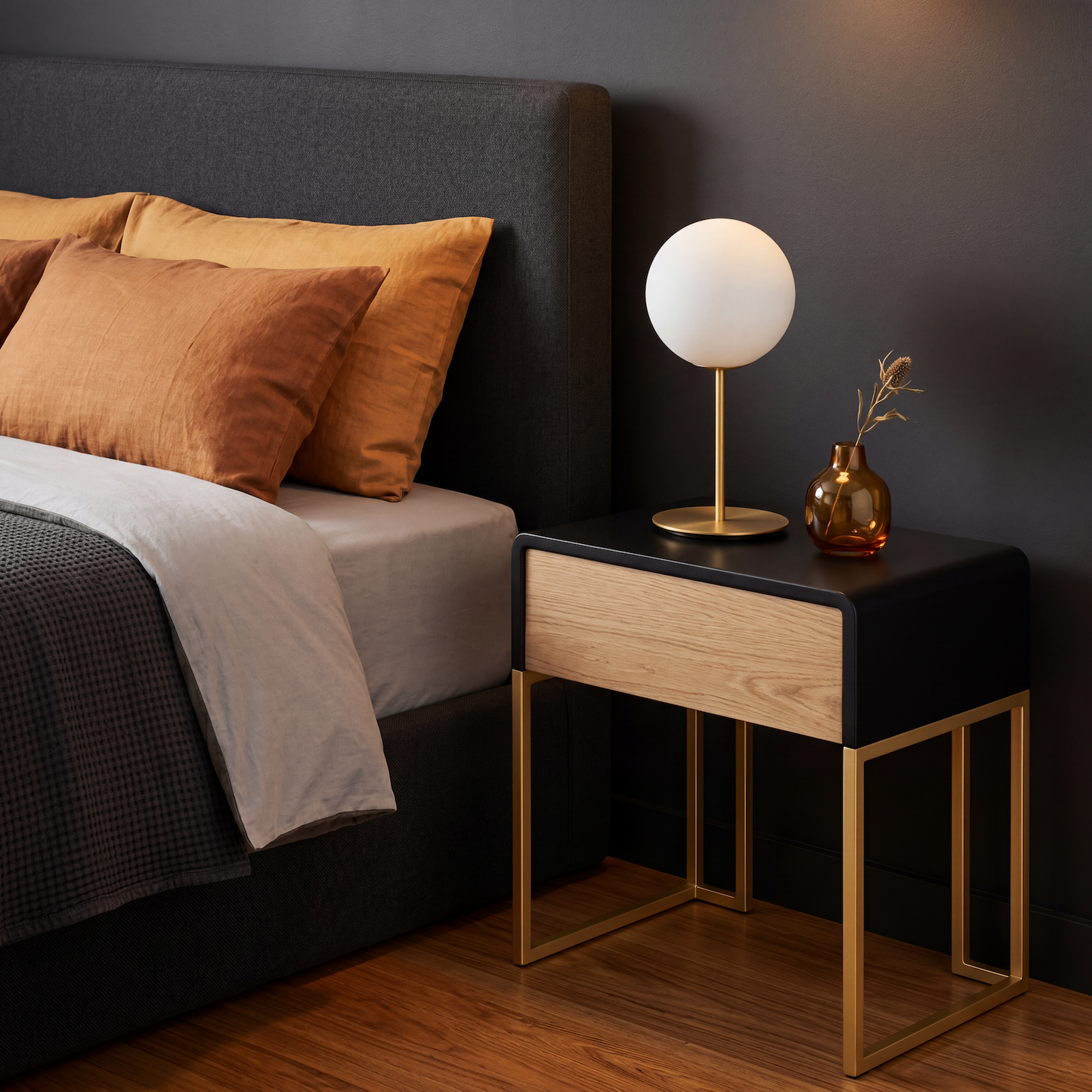 A modern bedside table in a bedroom setting, featuring a black top, a light oak wood drawer front, and gold metal legs. A white globe lamp and a small amber glass vase are styled on top next to a bed with a dark headboard and warm brown pillows.