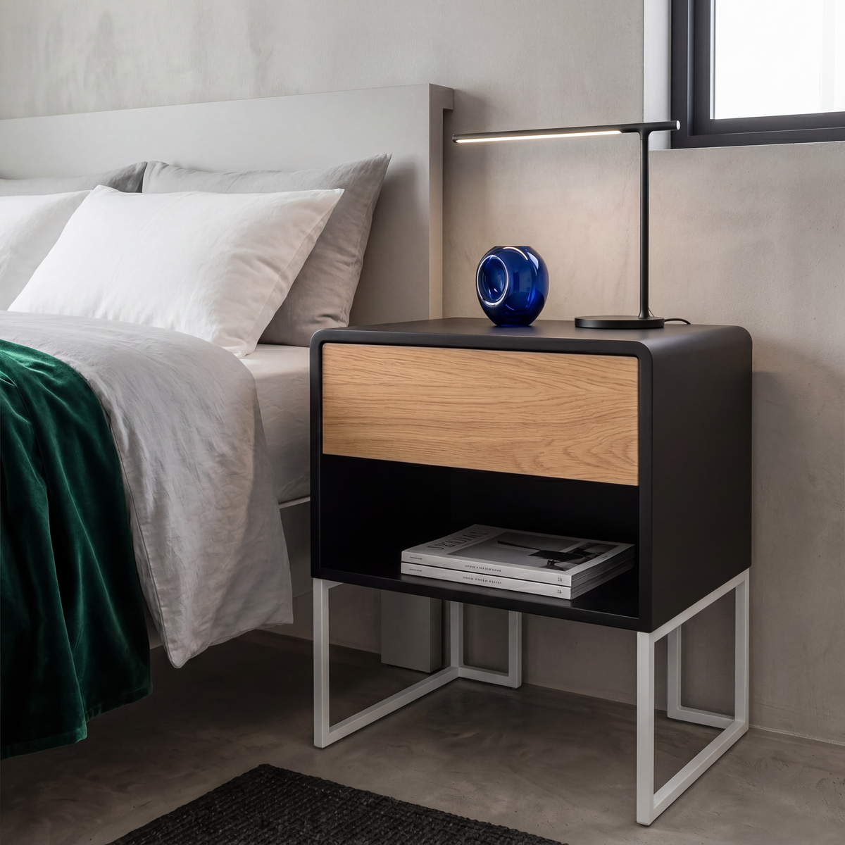 A modern black nightstand with a light wood drawer front and white metal legs, styled next to a bed in a minimalist bedroom setting.