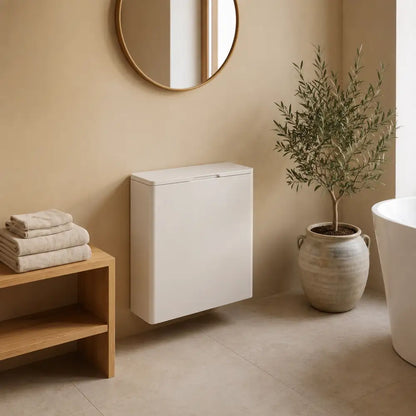 A wall-mounted white bathroom vanity cabinet in a warm beige bathroom setting, with folded towels on a wooden shelf, a potted olive tree, and part of a white bathtub visible.