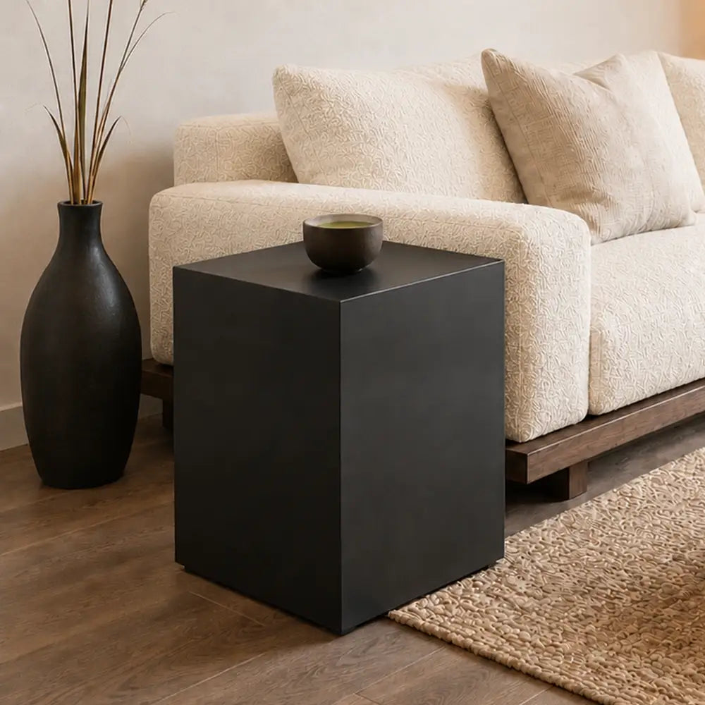 A matte black cube side table styled in a living room next to a cream boucle sofa, a tall black vase, and a woven rug.