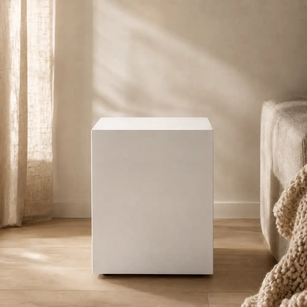 A minimalist white cube side table or stool in a warm, softly lit room with beige walls, light wood flooring, sheer curtains, and part of a sofa with a chunky knit throw visible on the right.