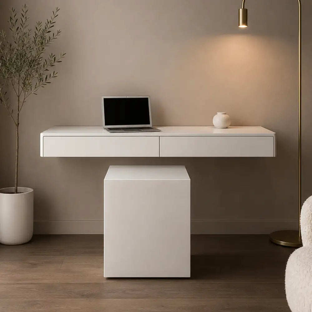 A modern white floating desk with two drawers and a matching cube-shaped storage ottoman, styled in a minimalist room with a laptop, small vase, floor lamp, and potted plant.