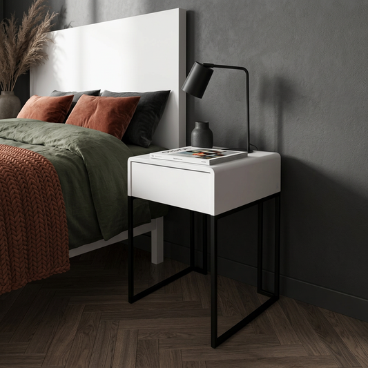 A modern white nightstand with a single drawer and open lower shelf, supported by black metal legs, styled in a bedroom next to a bed with green and rust bedding, a black table lamp, and a dark textured wall.
