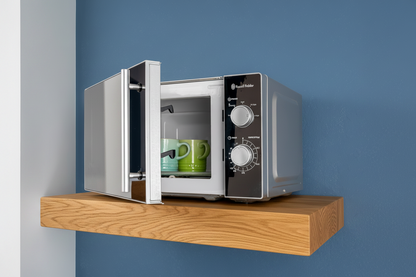 A compact countertop microwave oven with a silver exterior, black control panel, and open door showing green mugs inside, displayed on a wooden floating shelf against a blue wall.