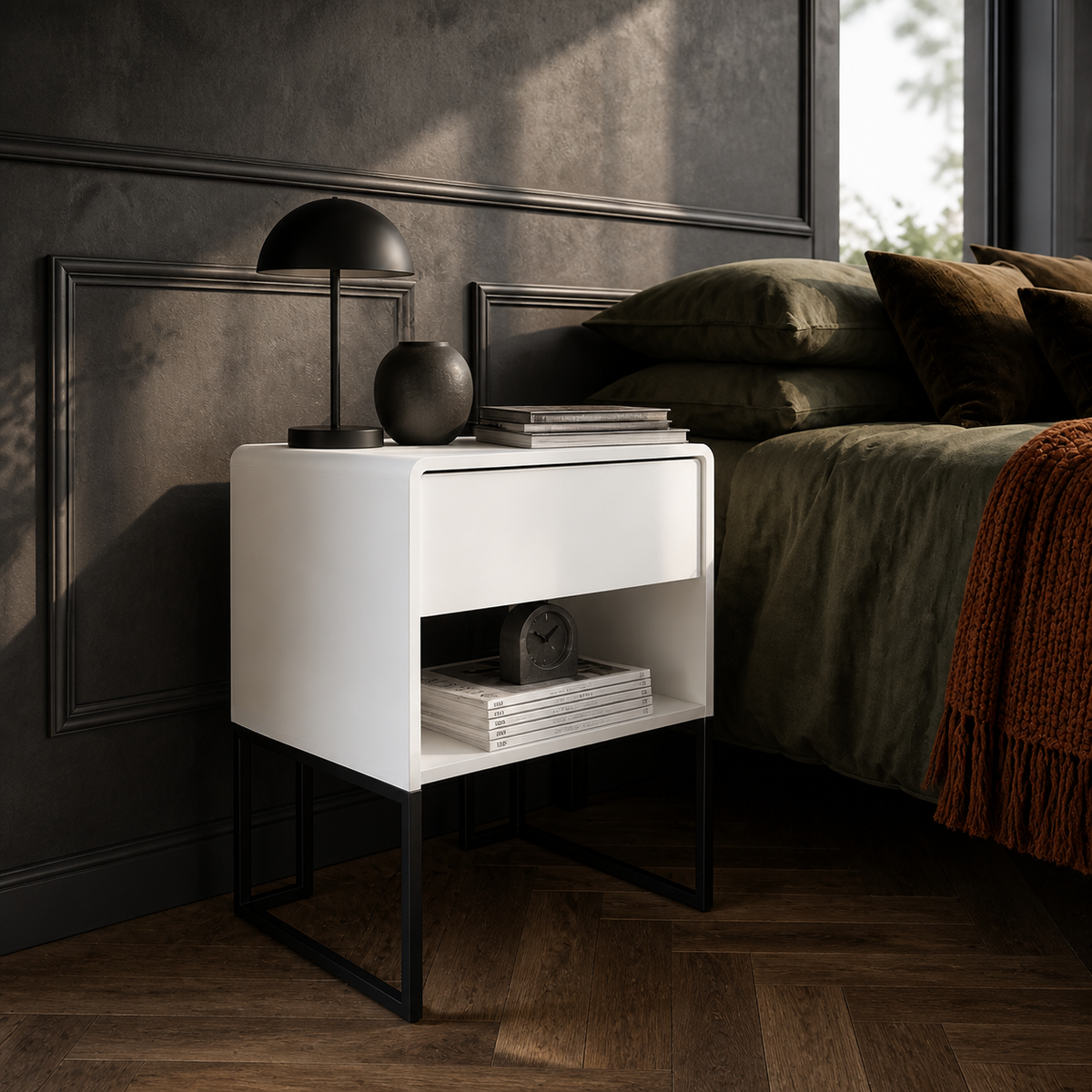 A modern white nightstand with an open lower shelf and black metal legs, styled in a dark bedroom next to a bed with olive green bedding and a black table lamp.