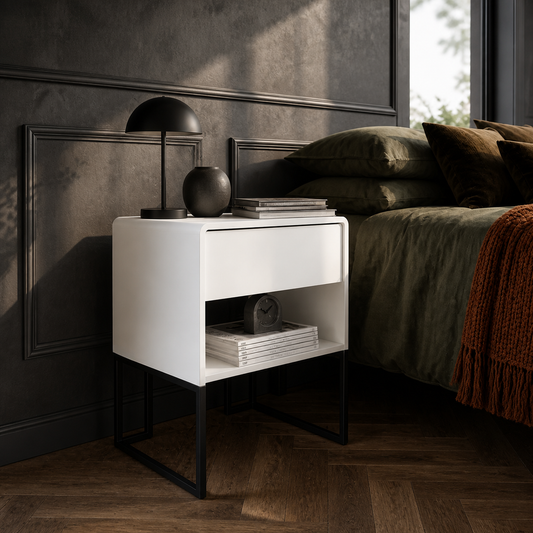 A modern white nightstand with an open lower shelf and black metal legs, styled in a dark bedroom next to a bed with olive green bedding and a black table lamp.