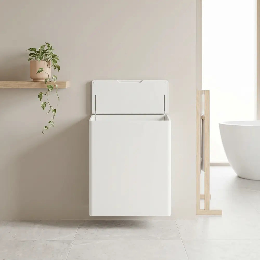 A minimalist white wall-mounted toilet paper holder with a hinged lid, shown in a clean, modern bathroom setting with a small wooden shelf and plant on the left.