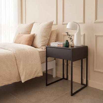 A modern bedside table with a dark charcoal gray top and black metal frame legs, styled in a bedroom next to a bed with beige linens and pillows.